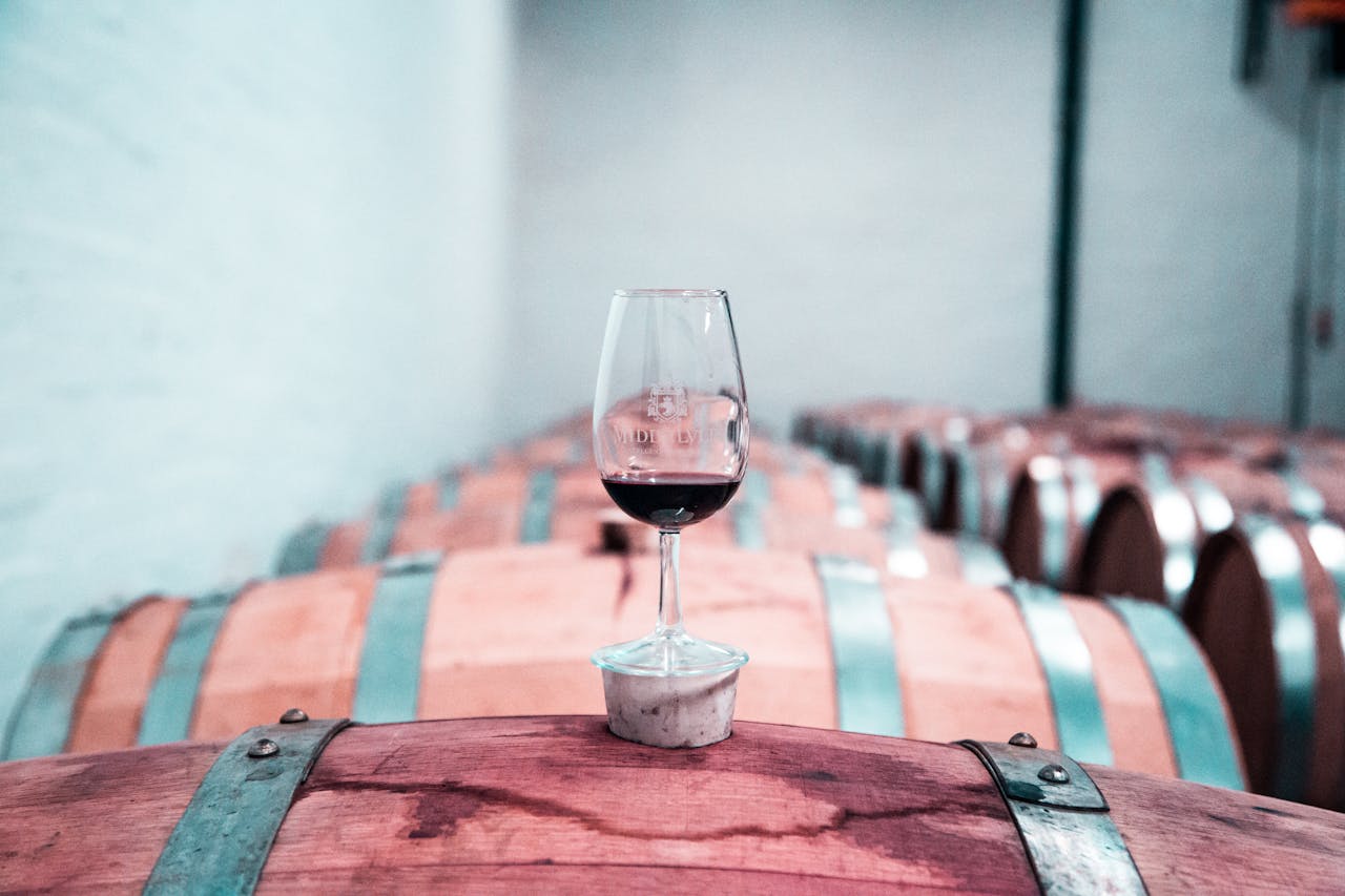 A glass of red wine sits elegantly on an oak barrel in a Stellenbosch winery cellar.